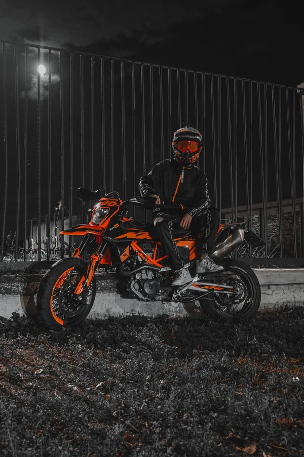 Un motociclista con casco accanto a una moto arancione, illuminati nella notte.