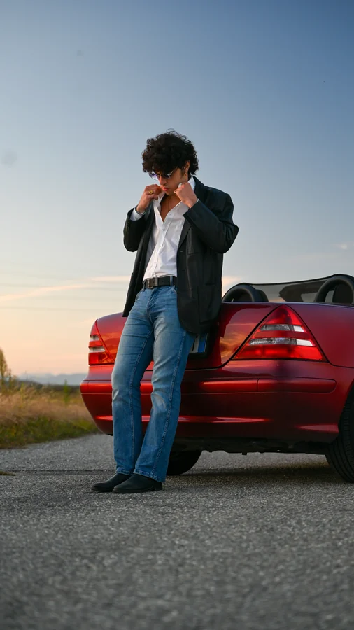 Una persona in giacca scura e jeans appoggiata a un’auto rossa al tramonto.